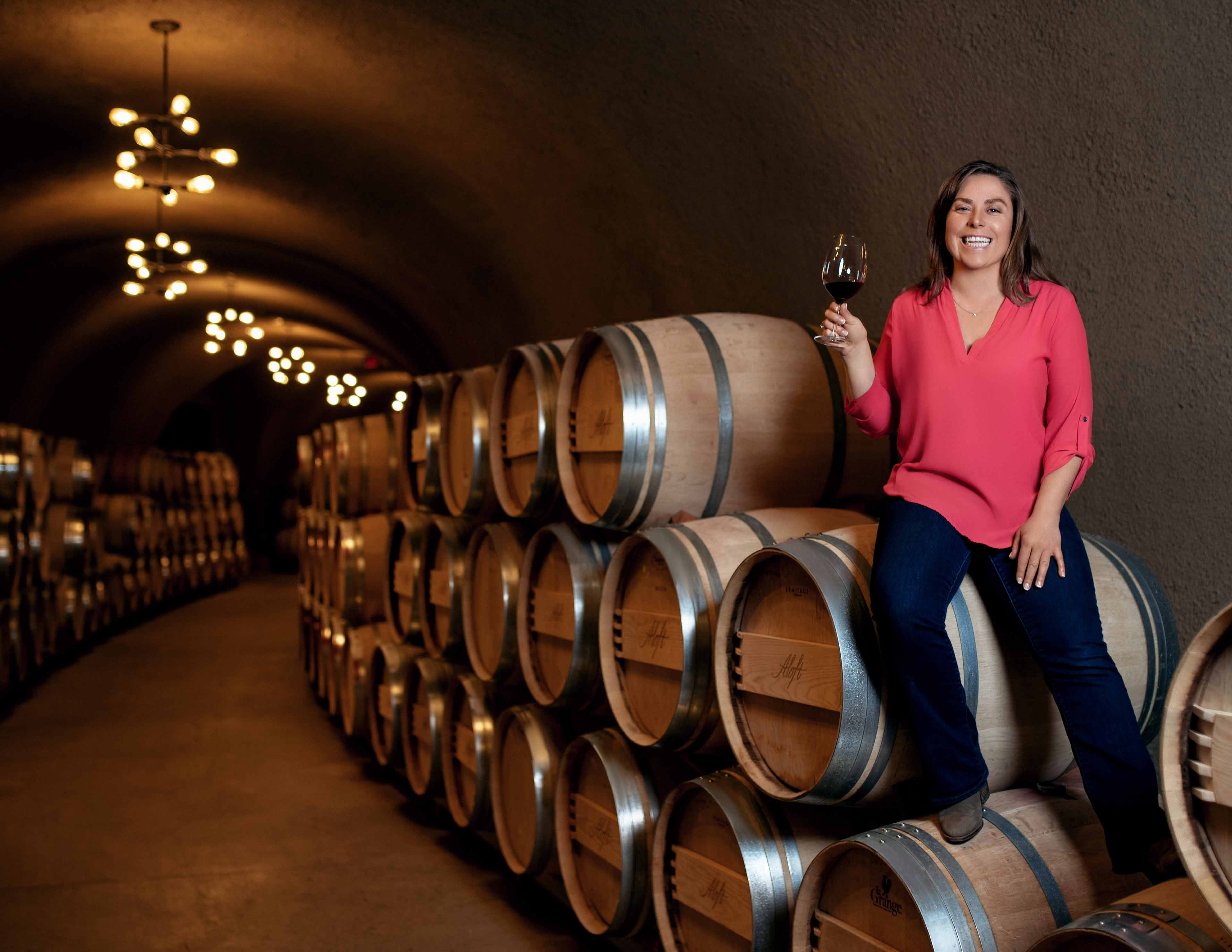 Angelina Mondavi sitting on some wine barrels holding a glass of wine in her hand