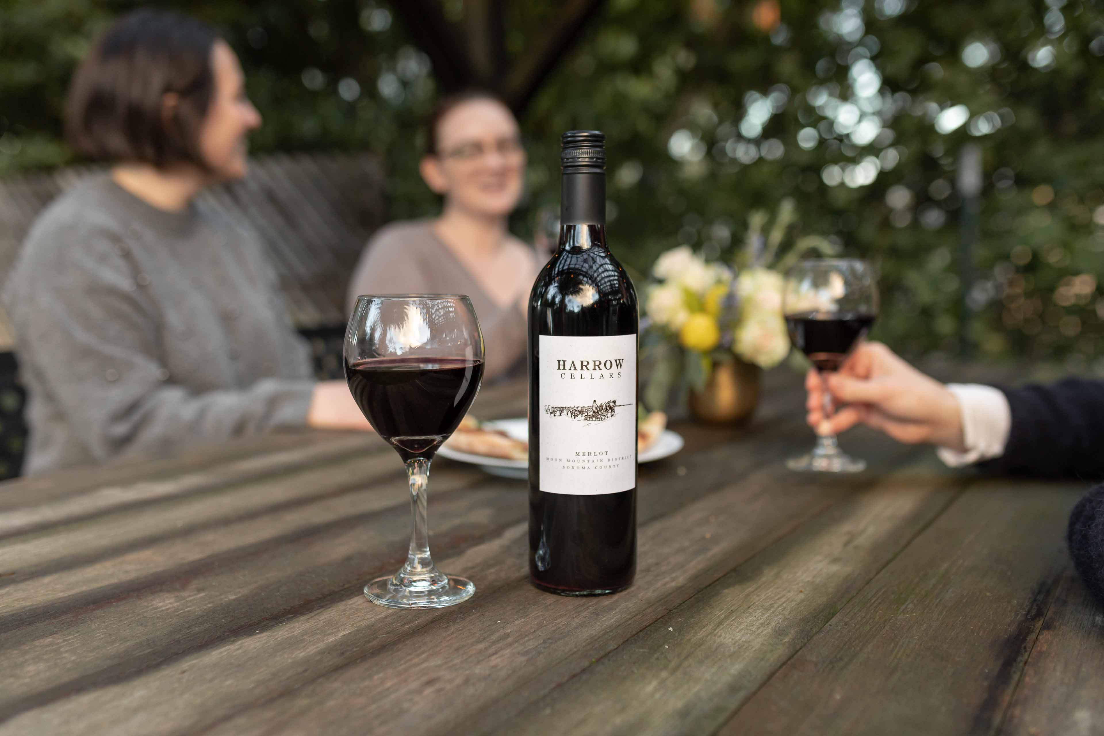 A Bottle Of Harrow Cellars Wine And A Glass Of Wine On A Table With People In The Background Eating And Drinking