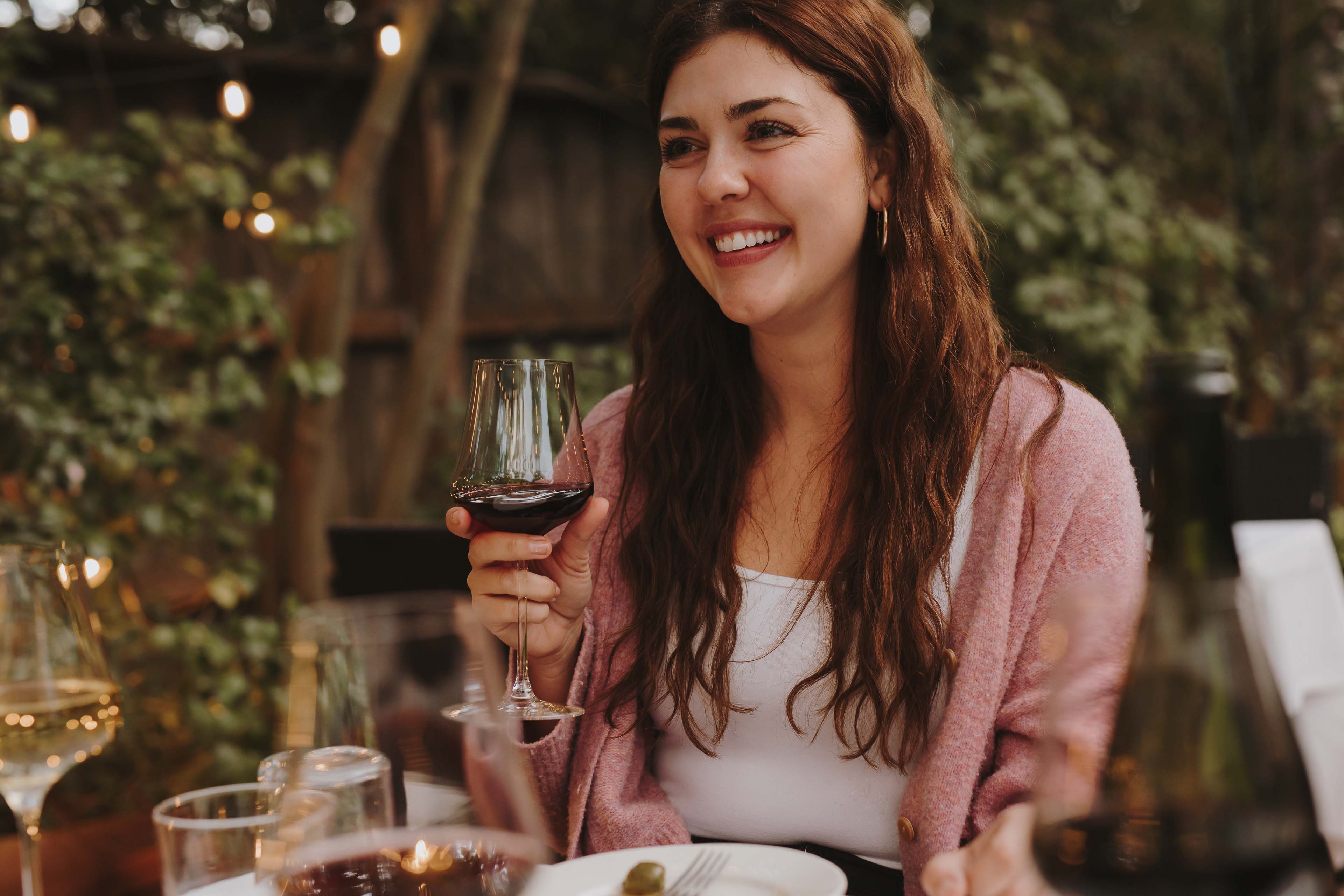 A Woman Sitting At An Outdoor Table Smiling With A Glass Of Red Wine In Hand